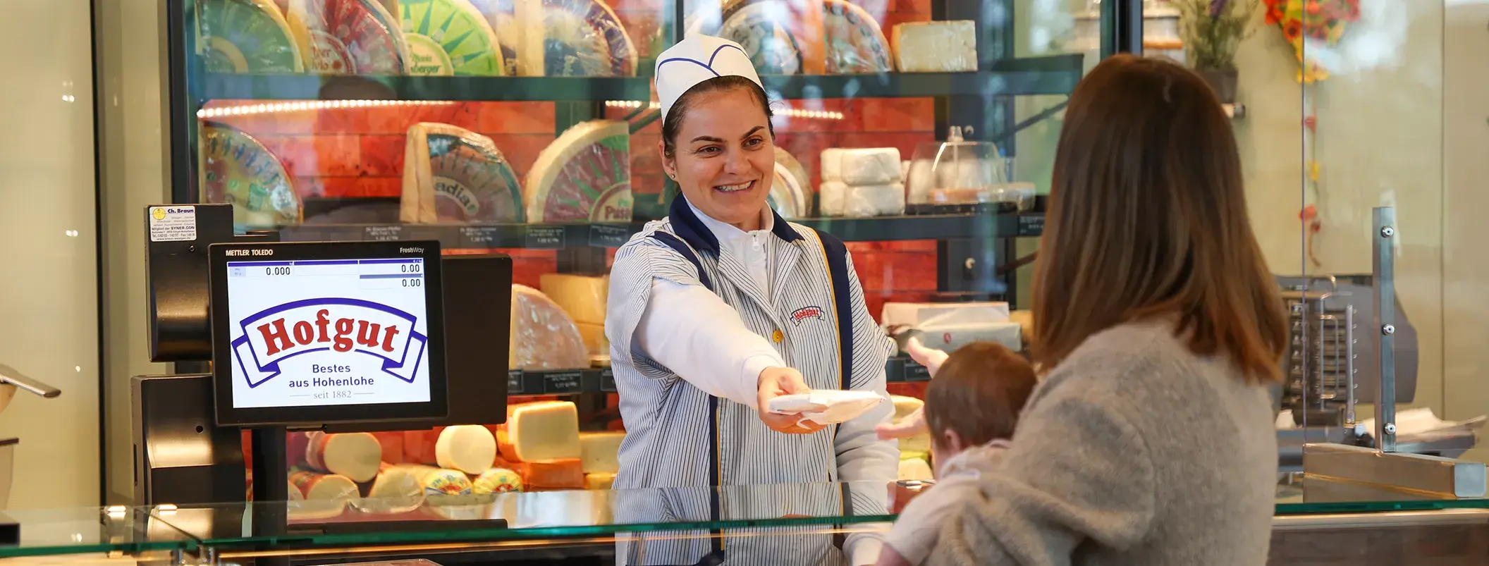 Eine Mitarbeiterin im Hofgut-Markt steht an der Theke und übergibt eingekaufte Lebensmittel an eine Kundin