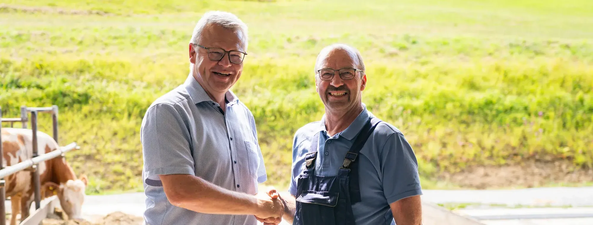 Der geschäftsführende Vorstand der Hohenloher Molkerei und ein Landwirt stehen händeschüttelnd im Stallausgang vor einer Wiese
