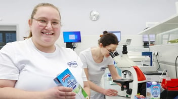 Viola Herrmann steht mit einer Packung Milch in der Hand im Labor und lächelt in die Kamera. Im Hintergrund arbeitet eine Kollegin am Mikroskop.