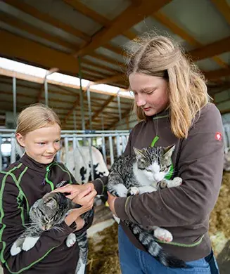 Zwei Töchter der Familie Riha stehen nebeneinander im Stall mit je einer Katze auf dem Arm