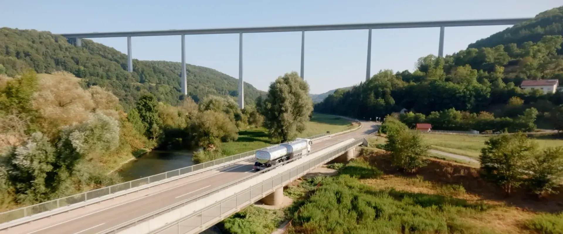 Ein Sammelwagen der Hohenloher Molkerei fährt auf einer Straße über eine Brücke, unter der ein Fluss entlang fließt durch eine grüne Landschaft