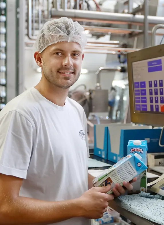 Ein Mitarbeiter der Hohenloher Molkerei steht in der Produktion vor einem Computer, lächelt in die Kamera und hält dabei eine Packung Milch in der Hand
