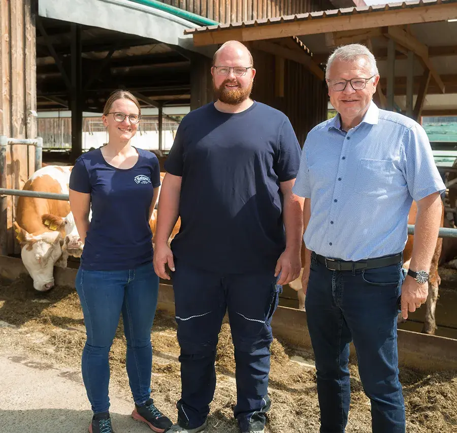 Die Leiterin der Abteilung Landwirtschaft, ein Landwirt und der geschäftsführende Vorstand der Hohenloher Molkerei auf dem Hof des Landwirts mit Kühen im Hintergrund.