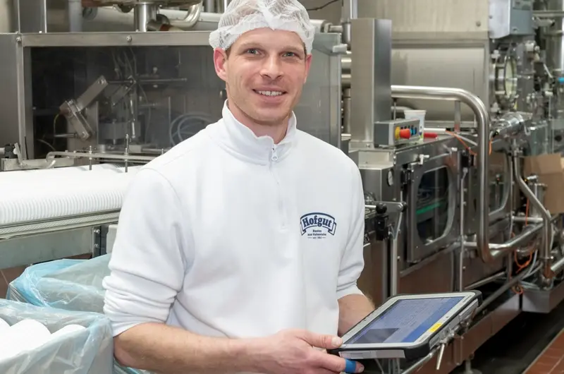 Mitarbeiter in Arbeitskleidung bedient Produktionsmaschine in der Molkerei und nutzt ein Tablet zur Qualitätssicherung.