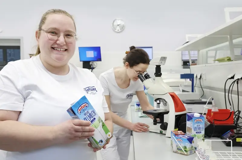 Zwei Laborantinnen prüfen Milch im modernen Labor der Hohenloher Molkerei – eine haelt eine Hofgut-Milch-Packung, die andere untersucht Qualität der Milch am Mikroskop.